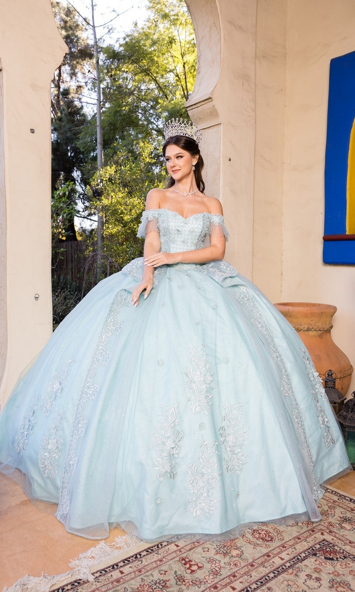 A young woman poses indoors near an arched doorway in the Quinceanera Dress 8055J by Cinderella Couture, an off-the-shoulder ball gown with a sweetheart neckline and subtle lace embroidery, greenery visible outside and a large vase beside her.
